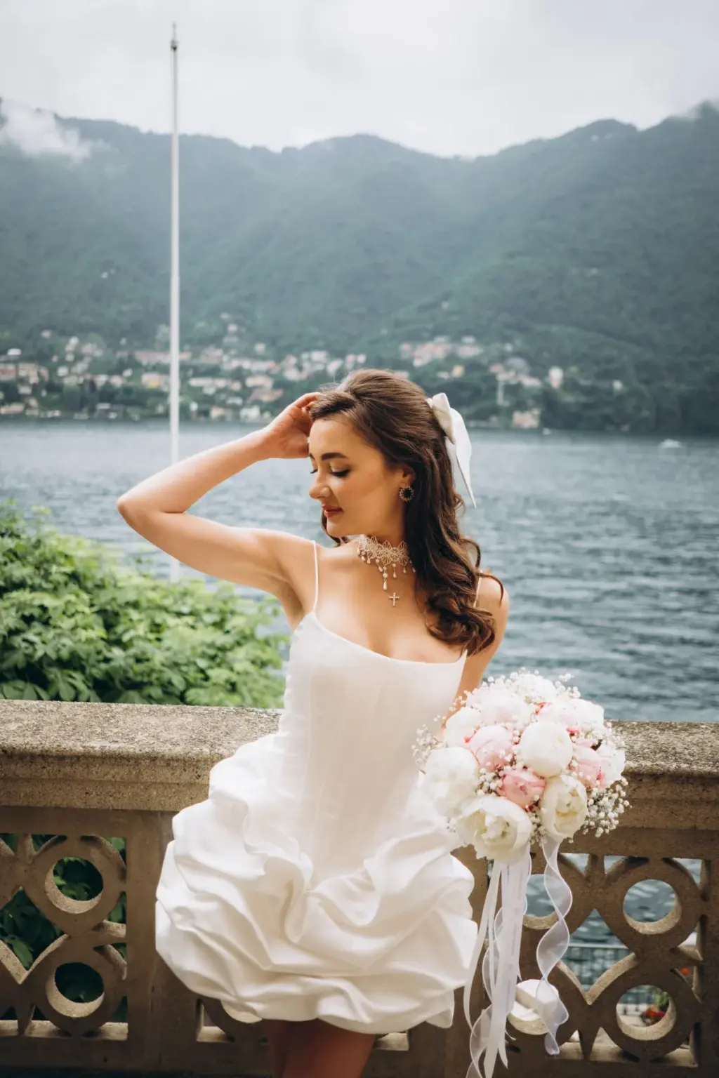 A bride touches her hair and turns her head on a lakeside balcony, a white bow in her hair and peony bouquet on the stone rail.