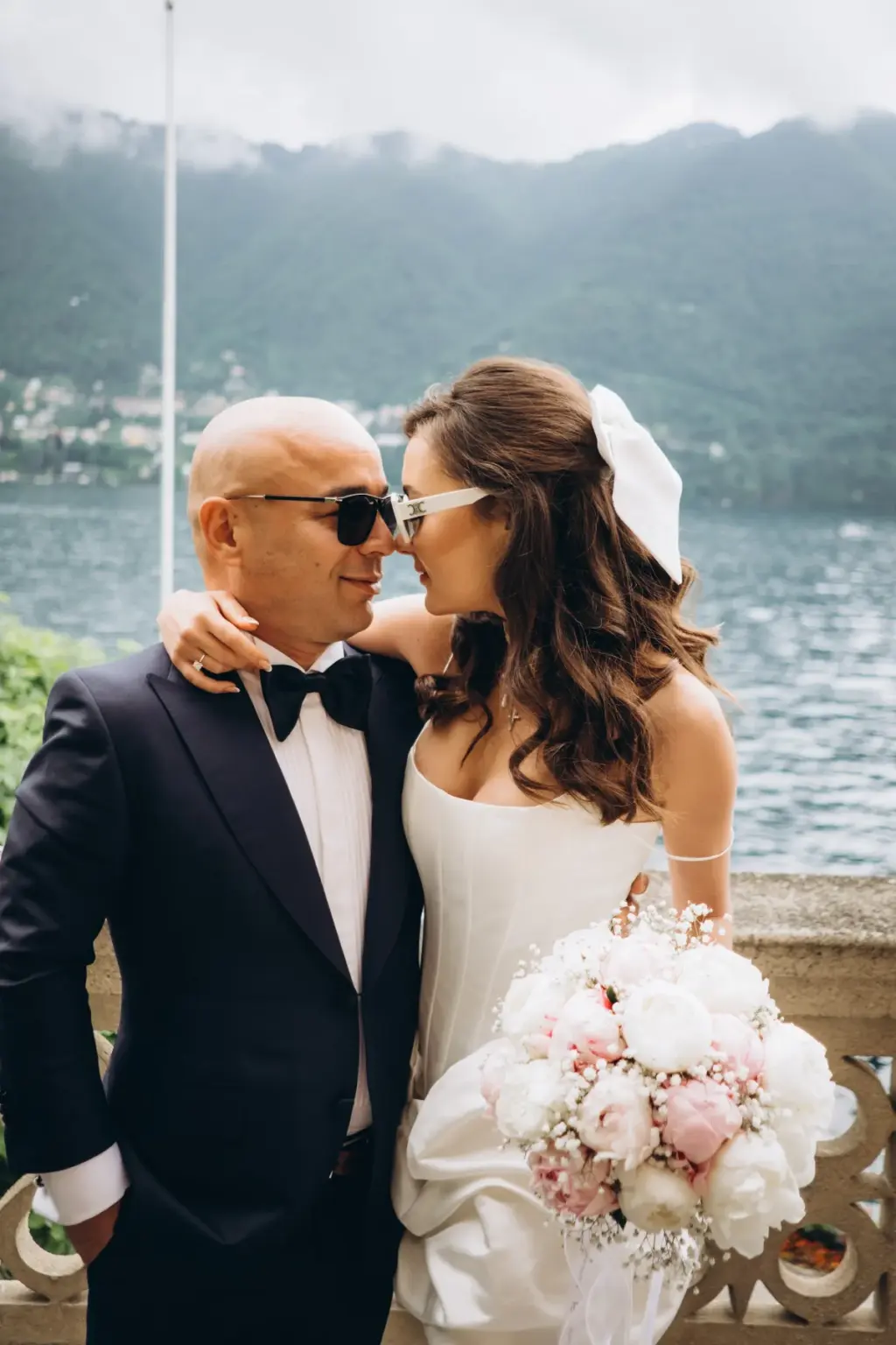 A bride and groom, both in dark sunglasses, smile nose to nose on a hotel balcony above Lake Como, her bouquet between them.