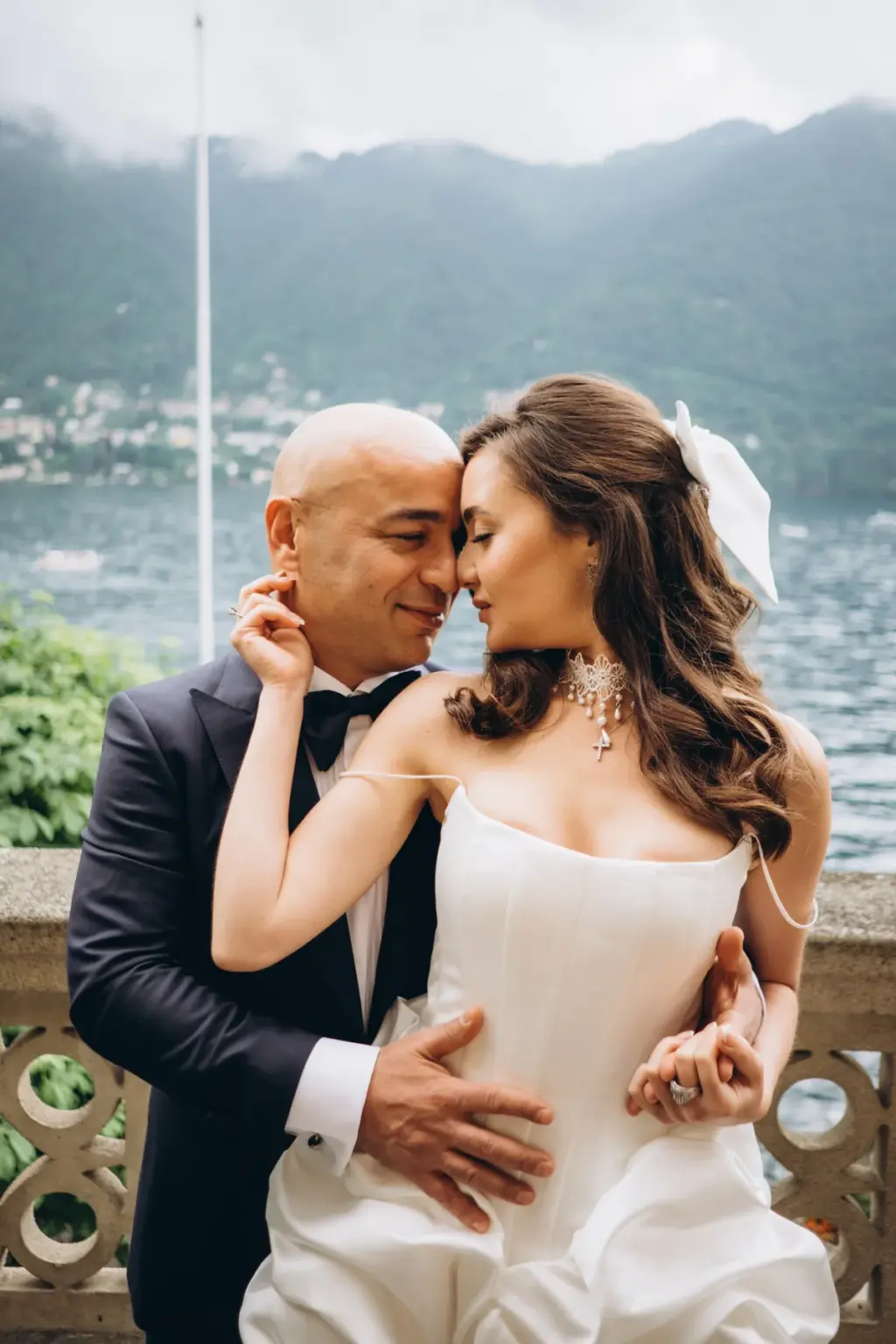 A bride with her hand on the groom's neck rests her forehead against his on a stone balcony, mountains and water behind.