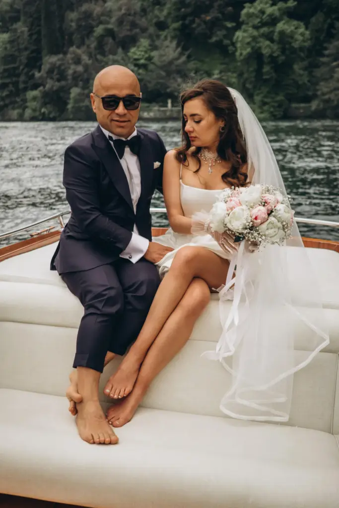A barefoot groom in a navy tuxedo sits beside the bride in her veil, her bouquet in her lap, on a boat with Lake Como behind.