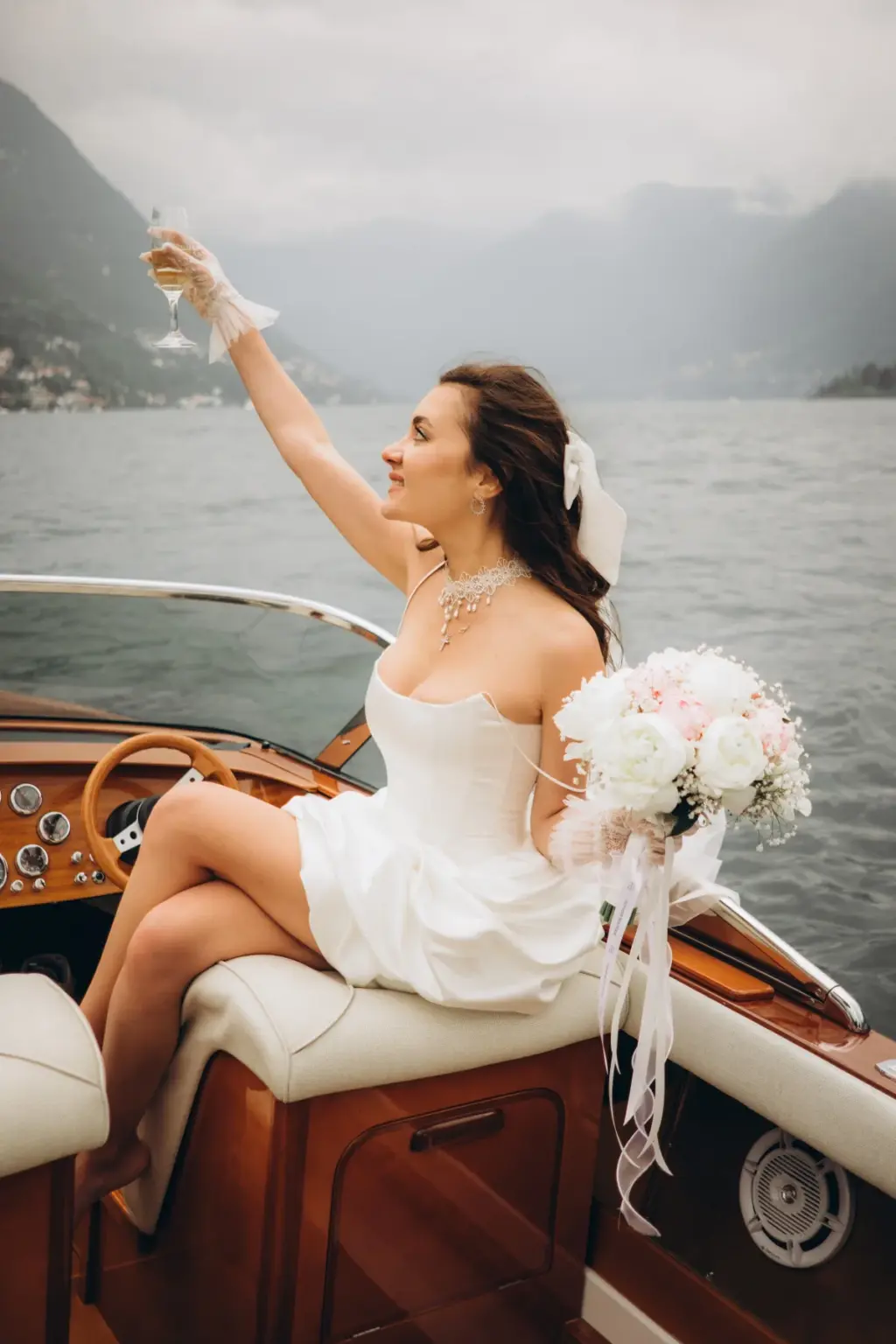 A bride in a short white dress and pearl necklace raises a glass of champagne high from the bow of a wooden boat on Lake Como.