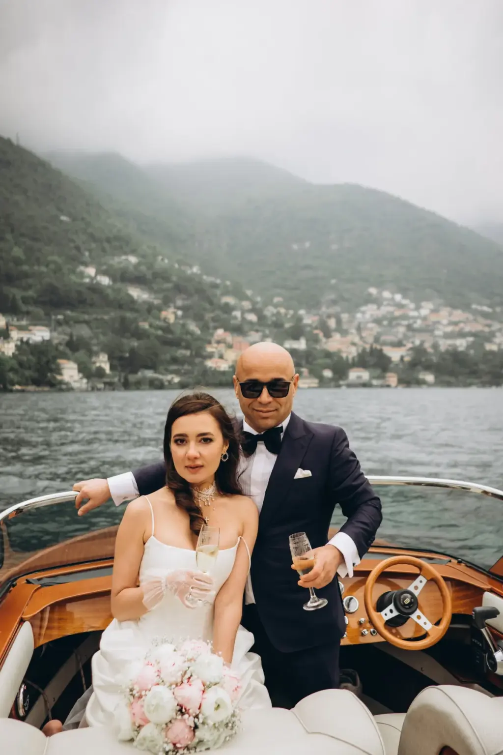 A bride with a champagne glass sits at the stern of a wooden boat while the groom stands behind her, misty mountains beyond.