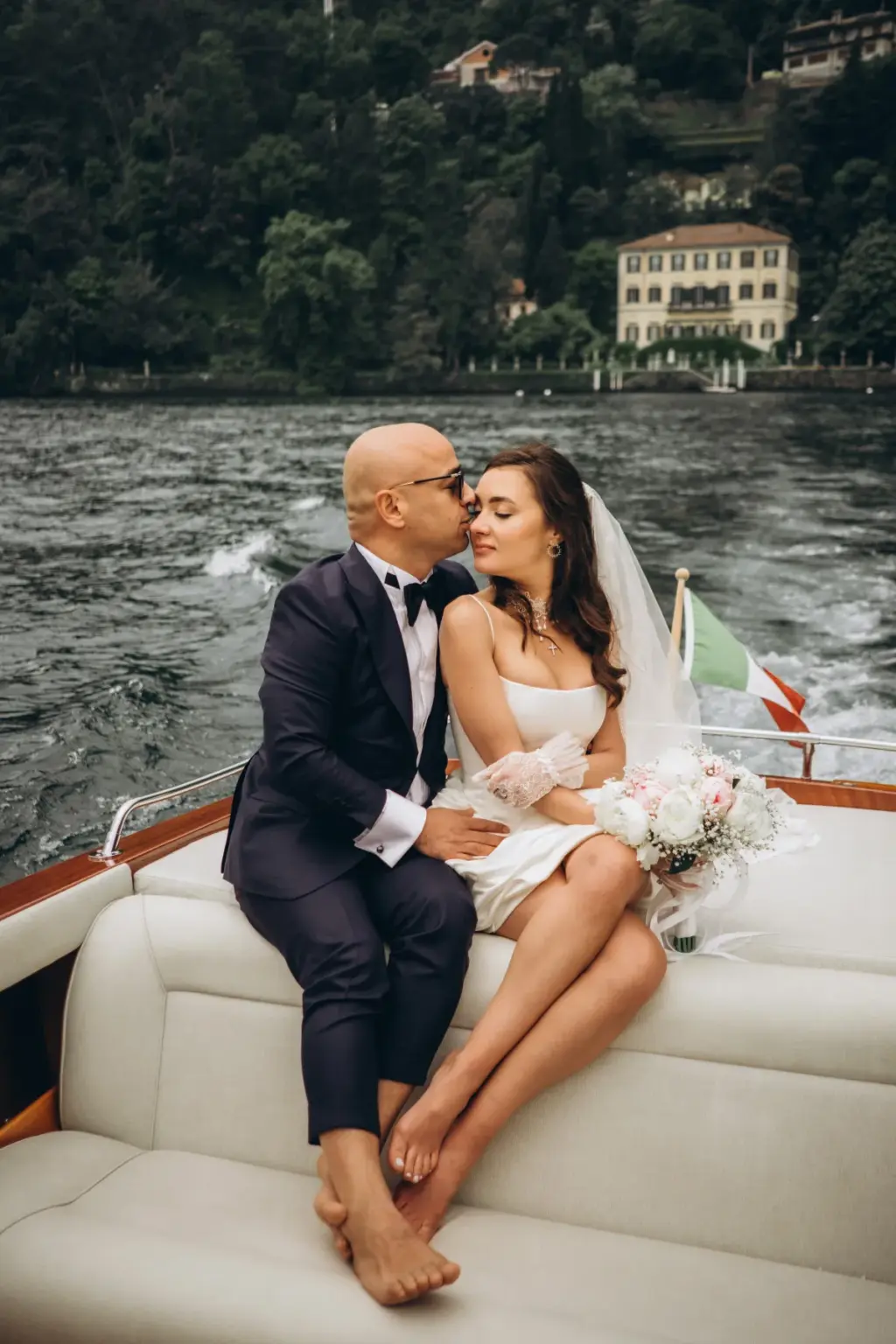 The groom leans in to kiss the bride's cheek on a wooden boat, a lakeside villa and green hillside rising behind them.