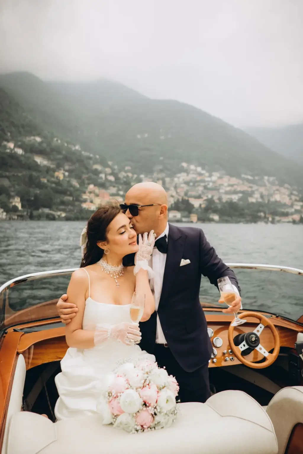 The groom kisses the bride's temple as she holds a glass of champagne in lace gloves on a boat, villages on the hills behind.