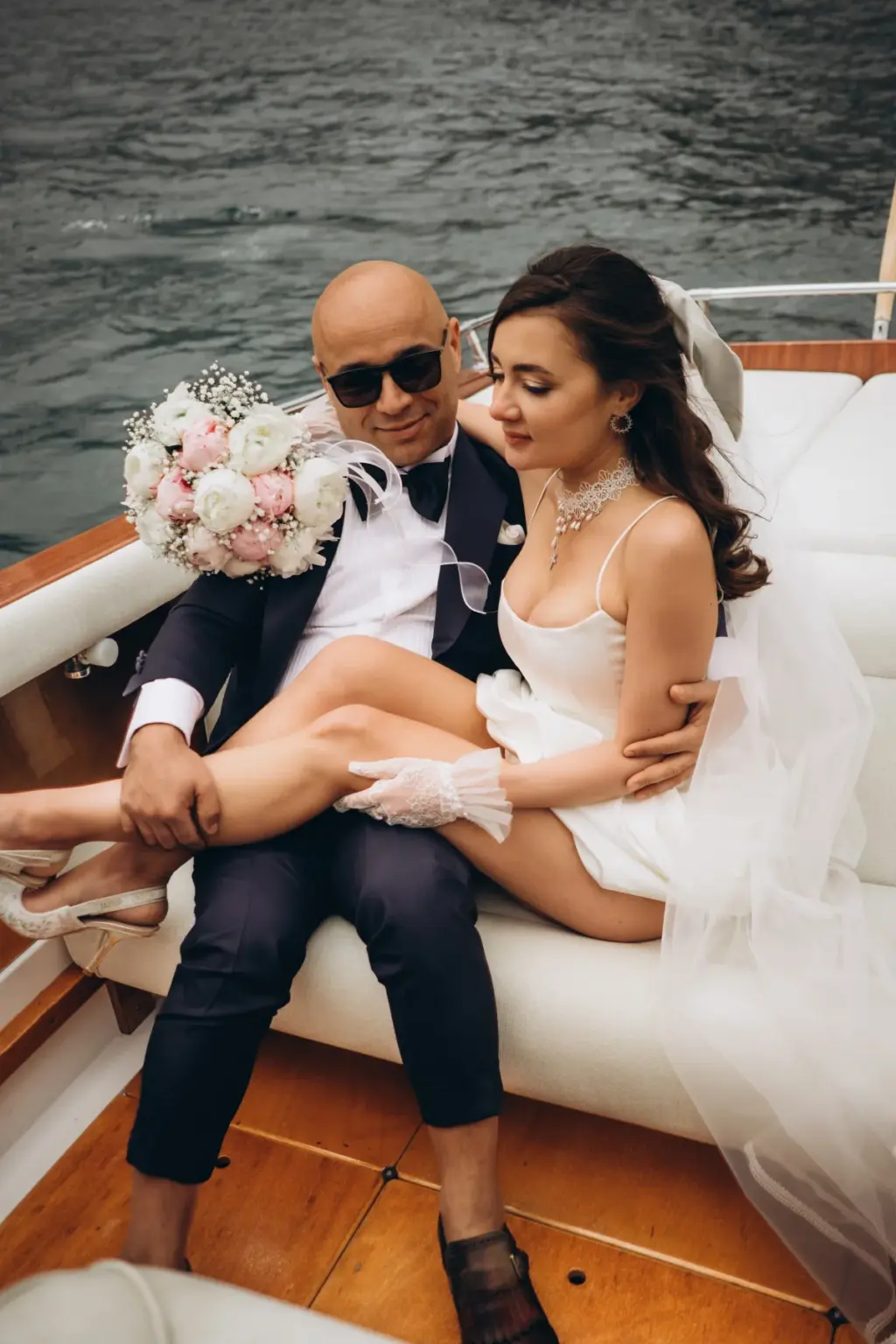 A groom in a tuxedo and sunglasses holds the bride close on the white leather seat of a boat, peony bouquet on the cushion behind.
