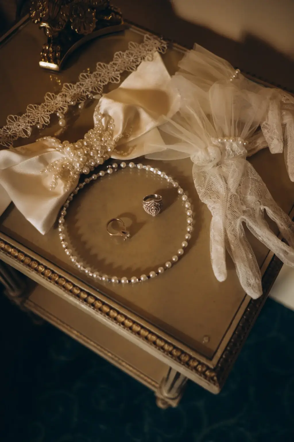 Wedding details on a gilded side table: a white satin hair bow, lace gloves, a pearl bracelet and two rings resting in its circle.