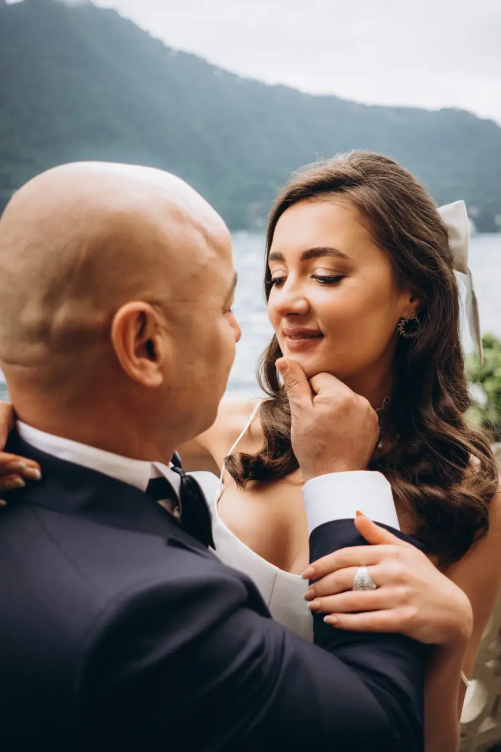 The groom gently lifts the bride's chin with his fingers as she smiles with eyes lowered, the green Lake Como shore behind them.