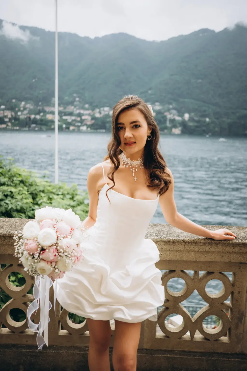 A bride in a short ruffled white dress and crystal necklace leans on a stone balcony rail, her bouquet set on it, Lake Como behind.