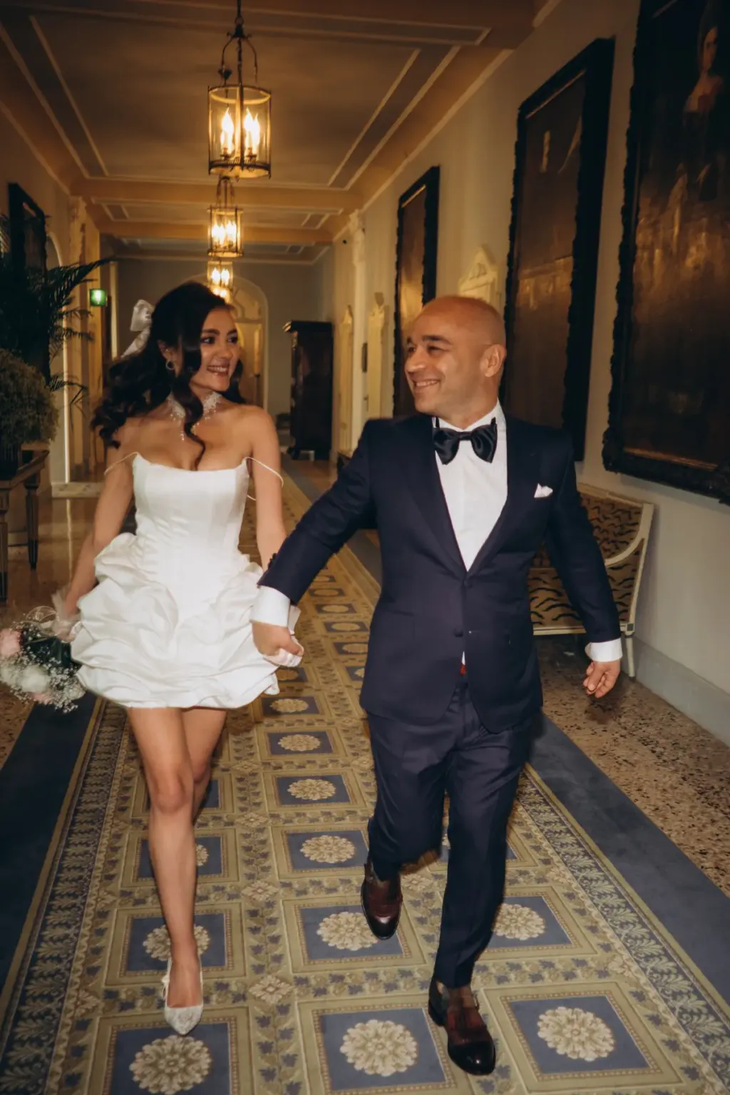 A laughing bride and groom stride hand in hand toward the camera along a hotel corridor with lanterns and a patterned carpet.