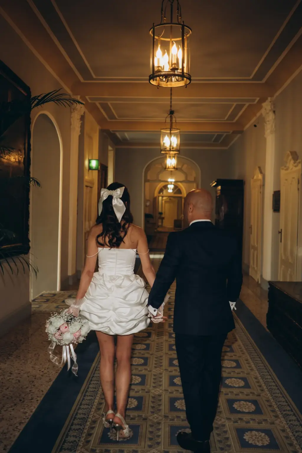 From behind, a bride with a white bow and a groom in a tuxedo walk hand in hand down a lantern-lit hotel corridor on a patterned rug.