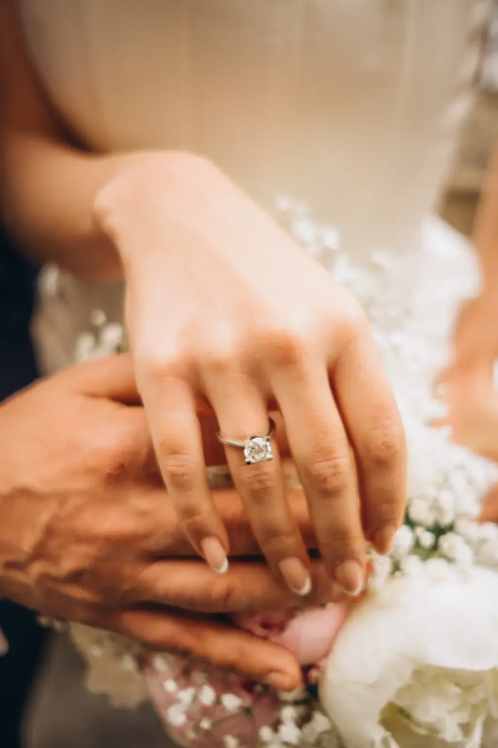 Close-up of the bride's hand with a large diamond engagement ring resting on the groom's hand over a bouquet of white flowers.