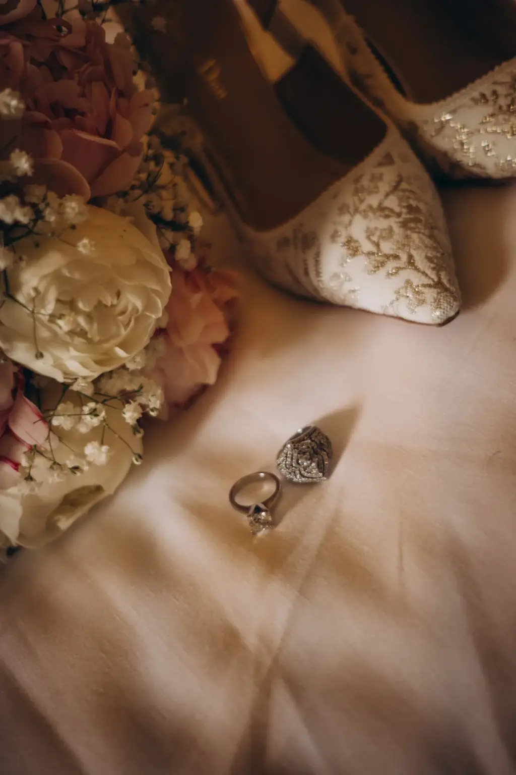 Two rings rest on a white bedspread beside a bouquet of white and pink roses and a pair of embroidered bridal heels.