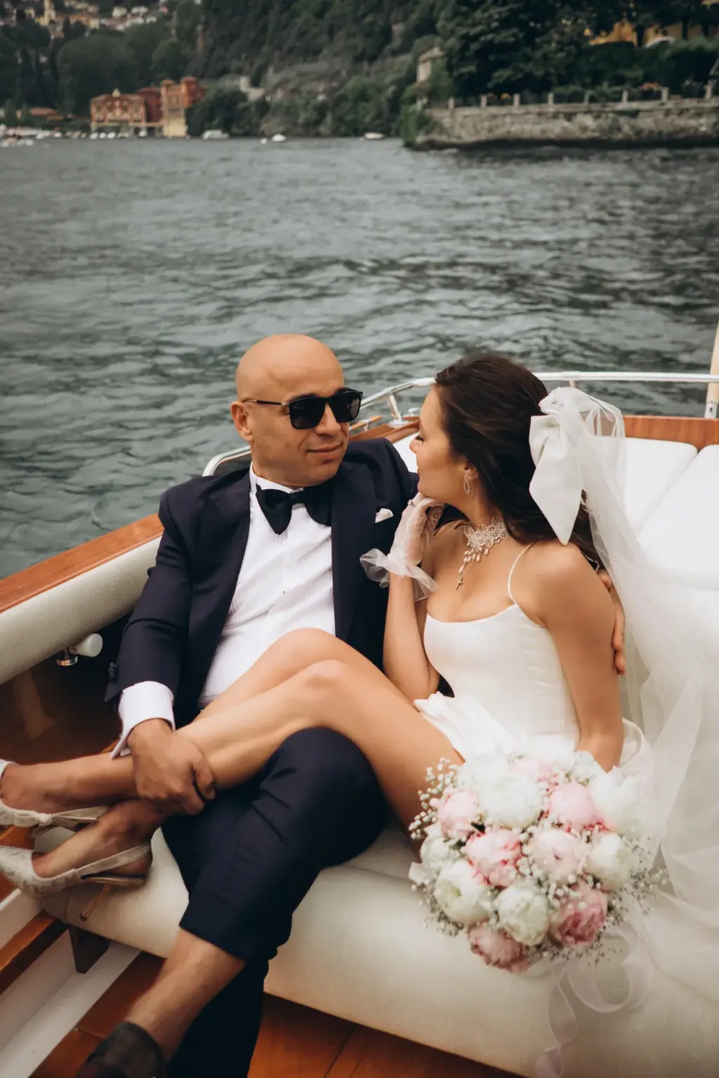 A bride in a short white dress and veil rests her legs across the groom's lap in a wooden boat, a pink and white bouquet beside her.