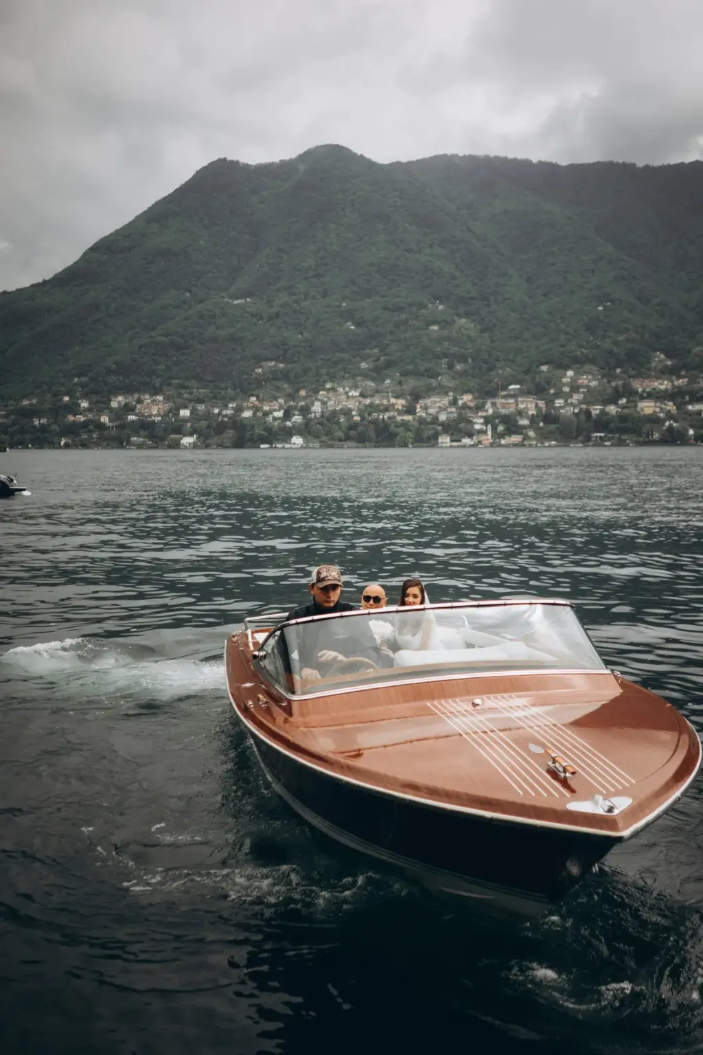 A varnished wooden Riva-style motorboat carries the bride, groom and driver across dark Lake Como water toward the camera.