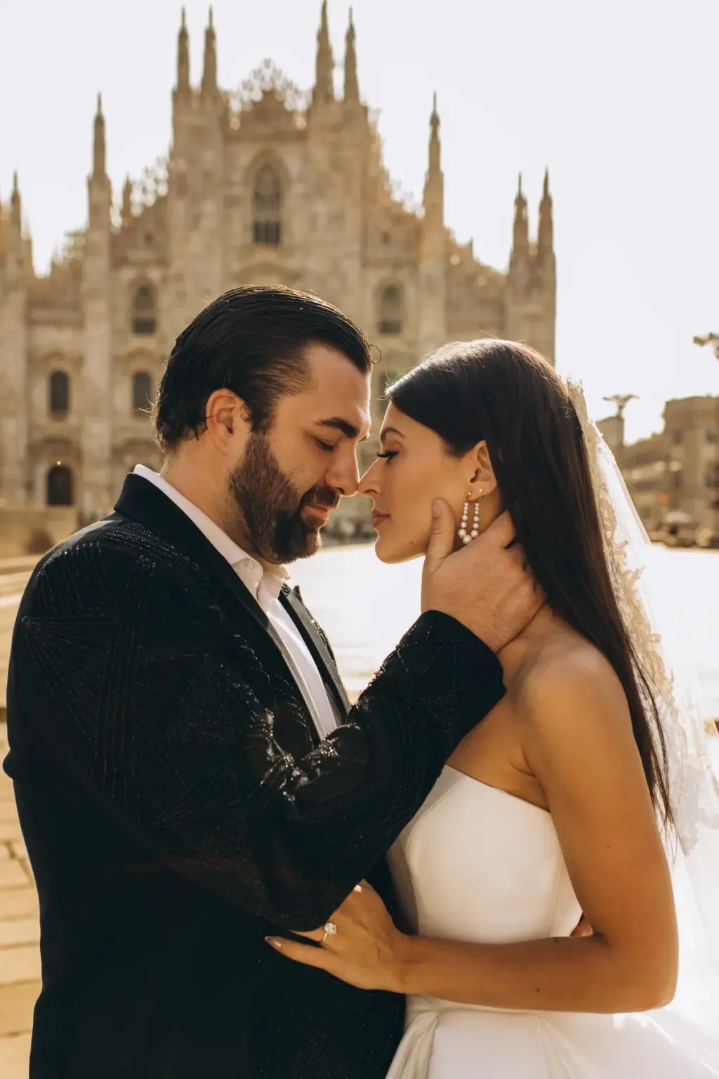 The bride cradles the groom's bearded face as they lean in close, the Duomo di Milano spires bright in the background.