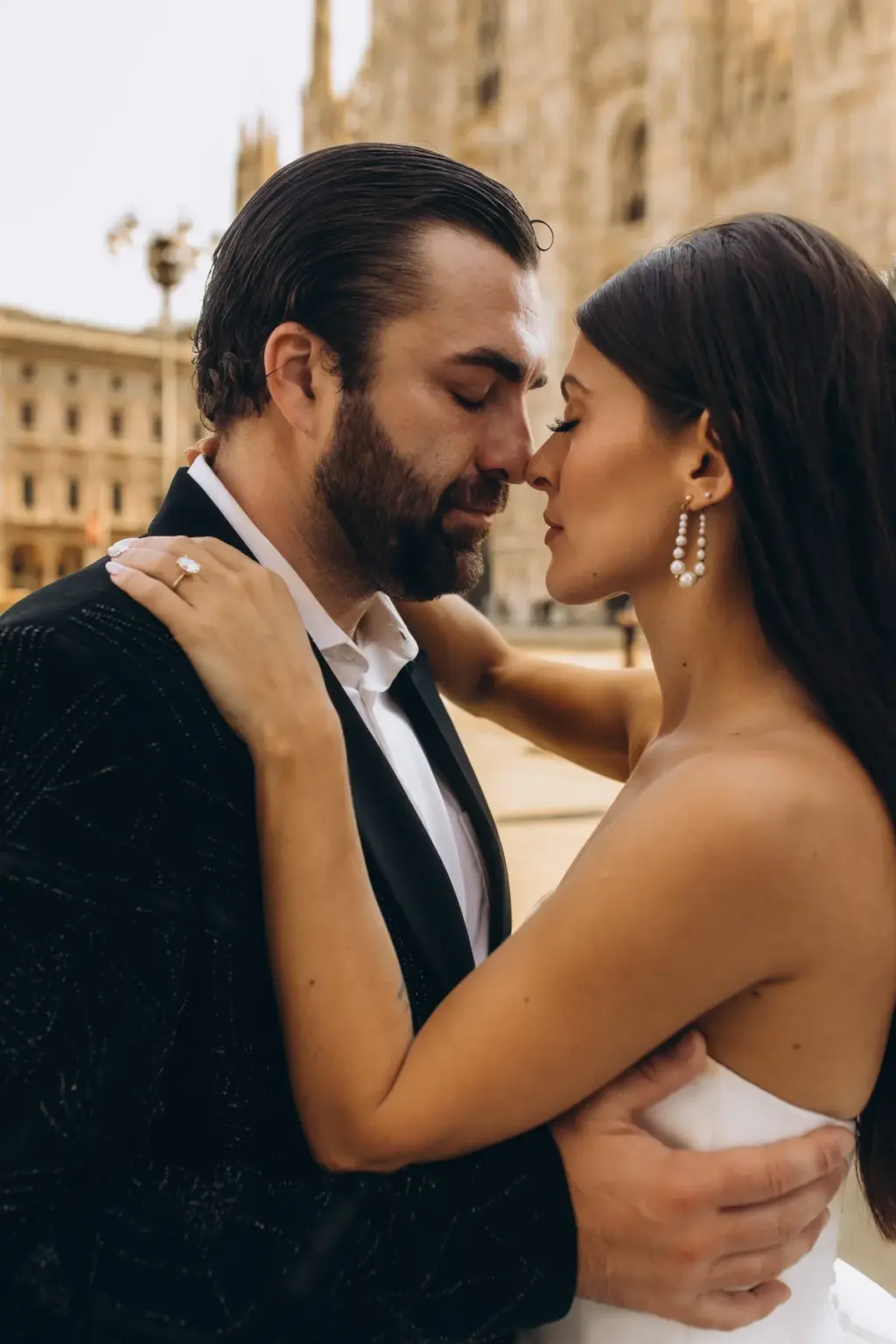 Close portrait of the bride and groom nose to nose with eyes closed, her ringed hand on his shoulder and drop earrings shining.
