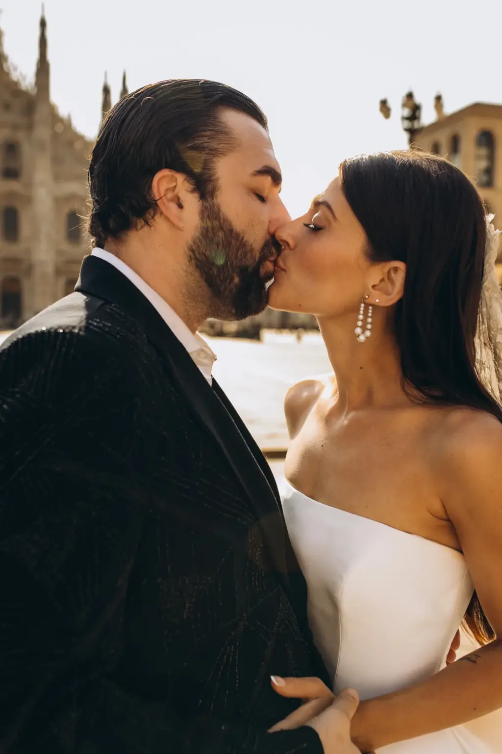 A tight close-up of the bride and groom kissing, her pearl drop earring and bare shoulders against his dark patterned jacket.
