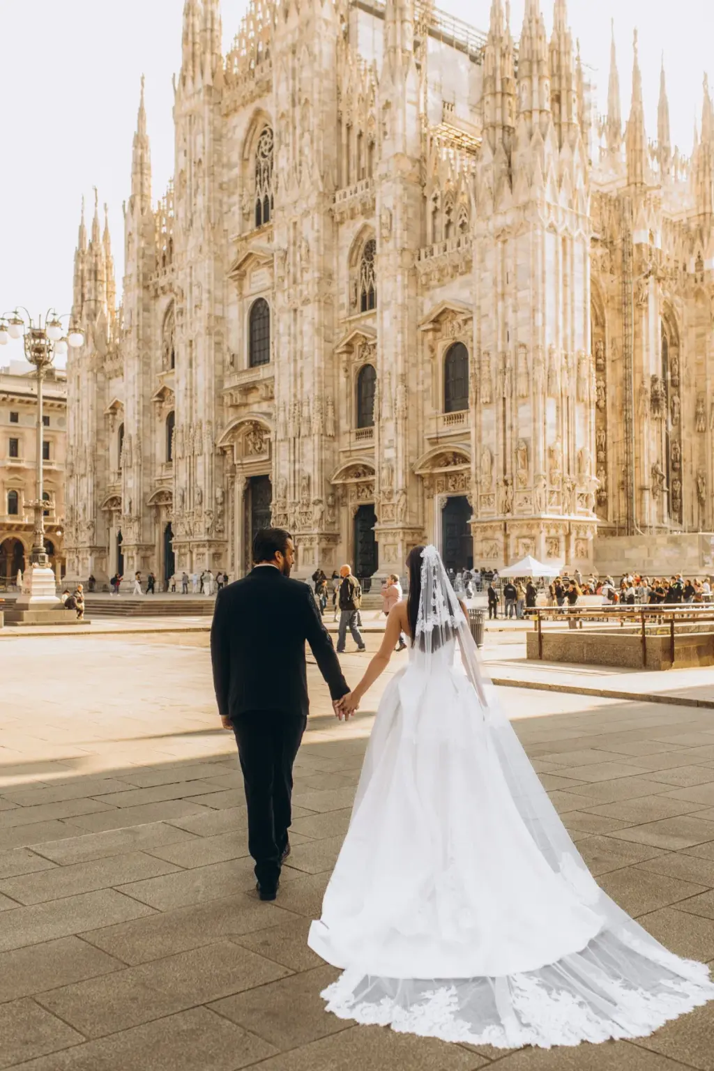 Seen from behind, the groom leads the bride by the hand toward the Duomo, her cathedral-length veil spilling over the stones.