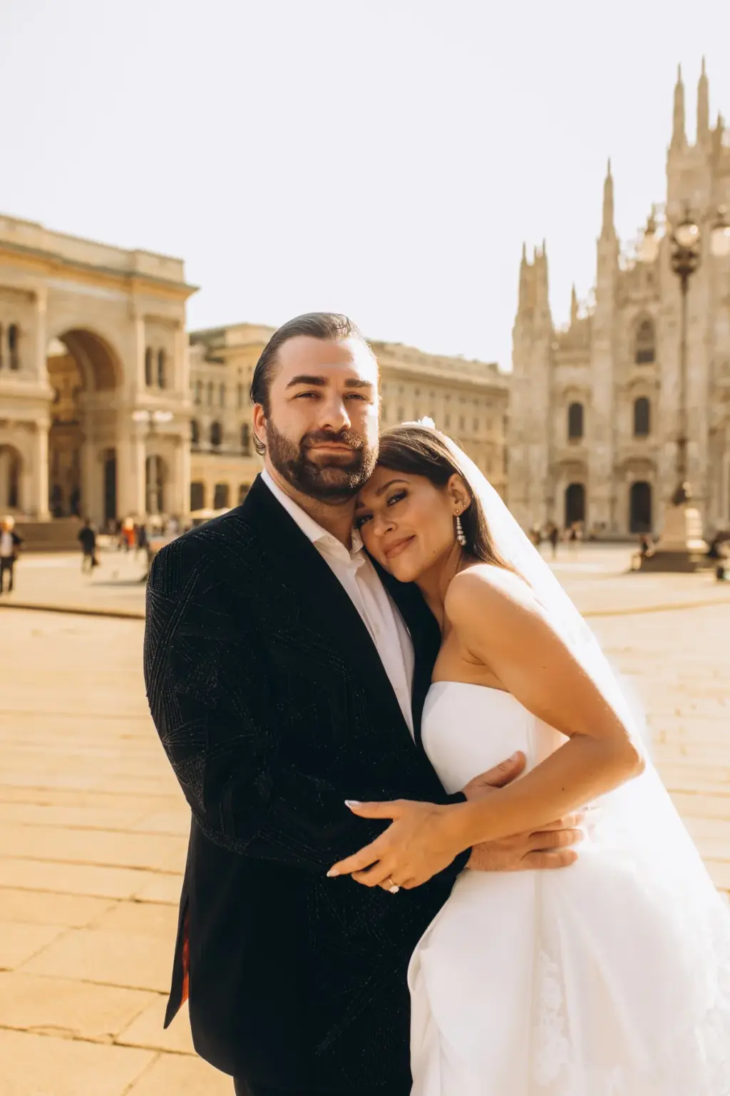 The bride rests her head on the groom's chest with eyes closed as he smiles at the camera, the Duomo and Galleria arch behind.