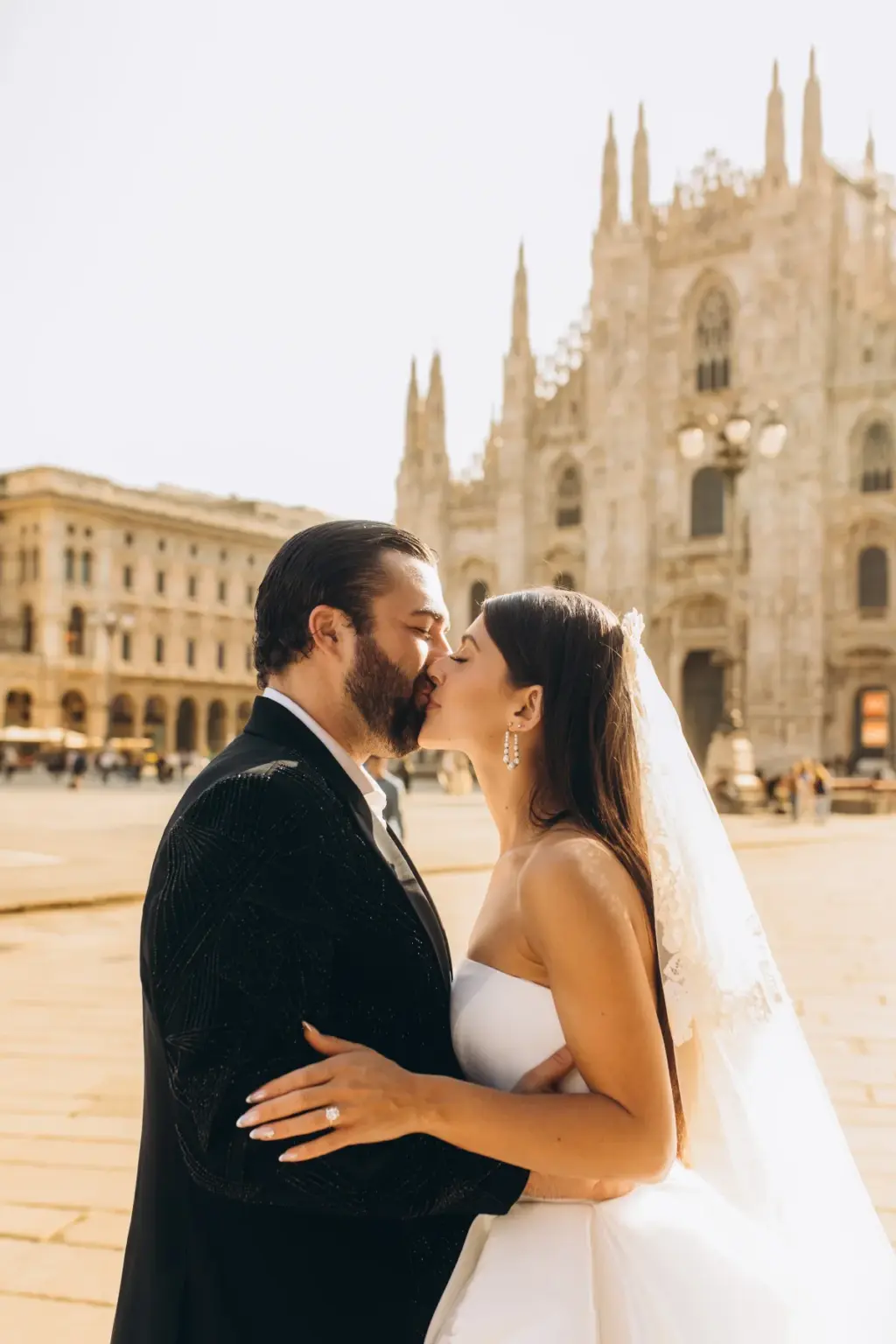 Bride and groom kiss on the Duomo square, her ring hand on his embroidered black jacket and her veil drifting behind.