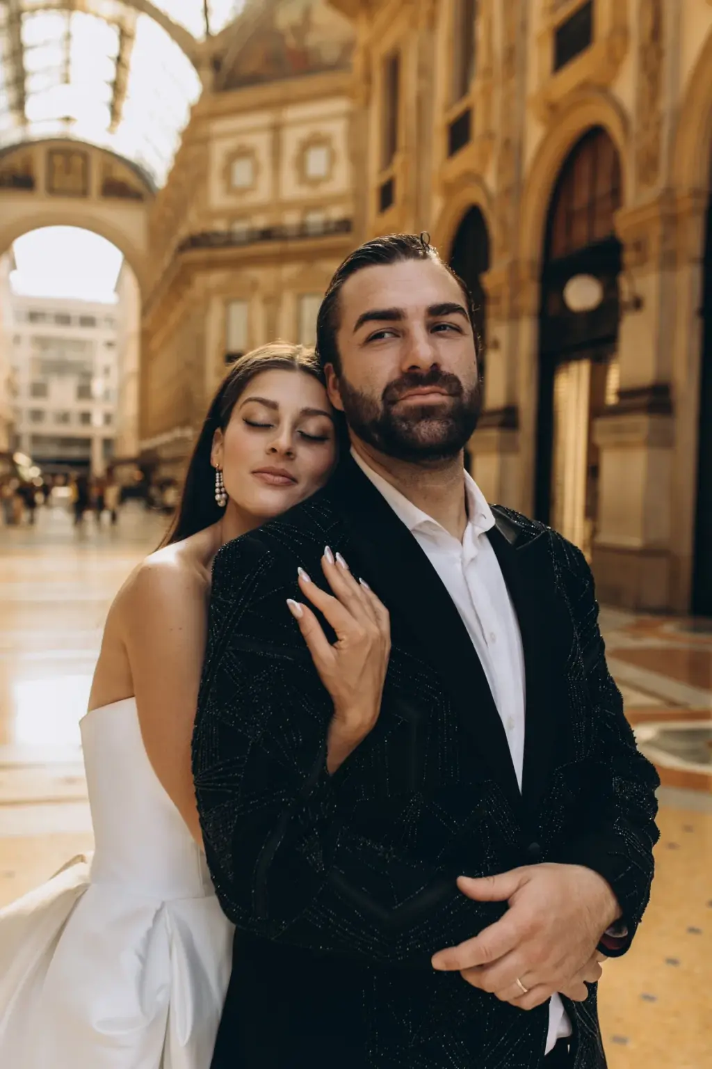 Standing behind the groom, the bride rests her cheek on his shoulder with eyes closed while he looks into the camera in the Galleria.