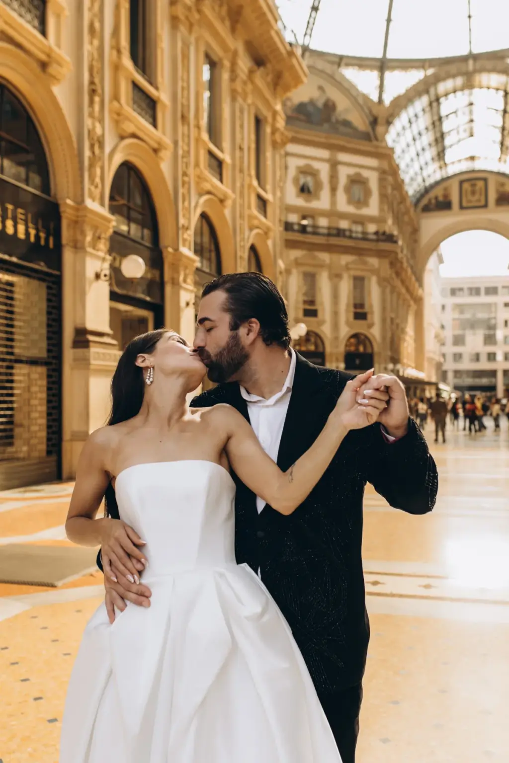 In a waltz hold, the bride rises onto her toes to kiss the groom inside the Galleria, its glass arcade glowing behind.