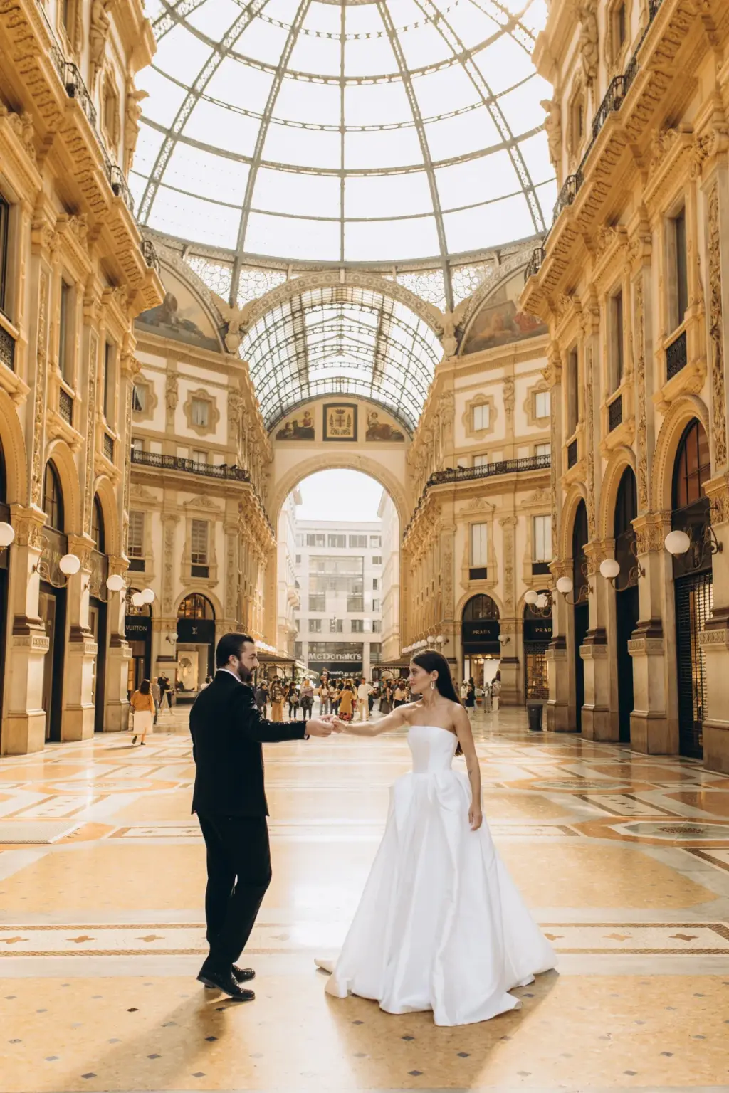 Bride and groom reach for each other's hands at arm's length beneath the glass dome of Galleria Vittorio Emanuele II.