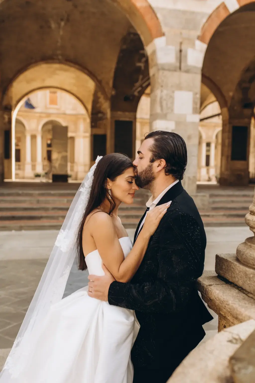 The bride leans into the groom with eyes closed as he holds her waist, framed by the vaulted arcade and its worn stone steps.