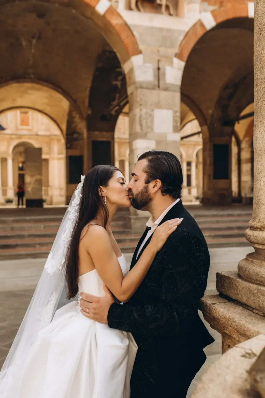 Beside a stone column under the brick arches of Piazza dei Mercanti, the bride kisses the groom, her veil falling to the ground.