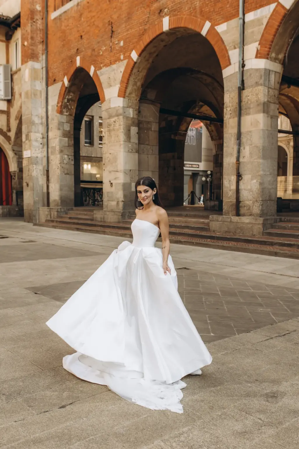 The bride swirls her full satin skirt alone on the paving of Piazza dei Mercanti, the red-brick arches and steps behind her.