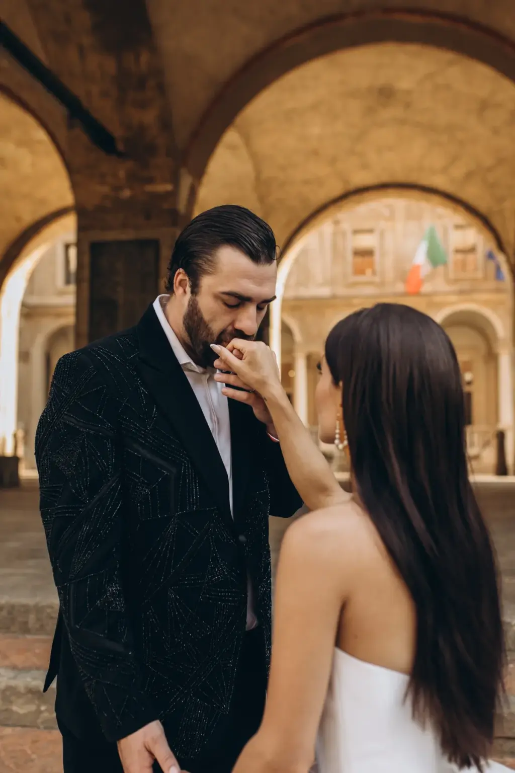 The groom kisses the bride's hand under a vaulted arcade, an Italian flag hanging in the courtyard seen through the arches.