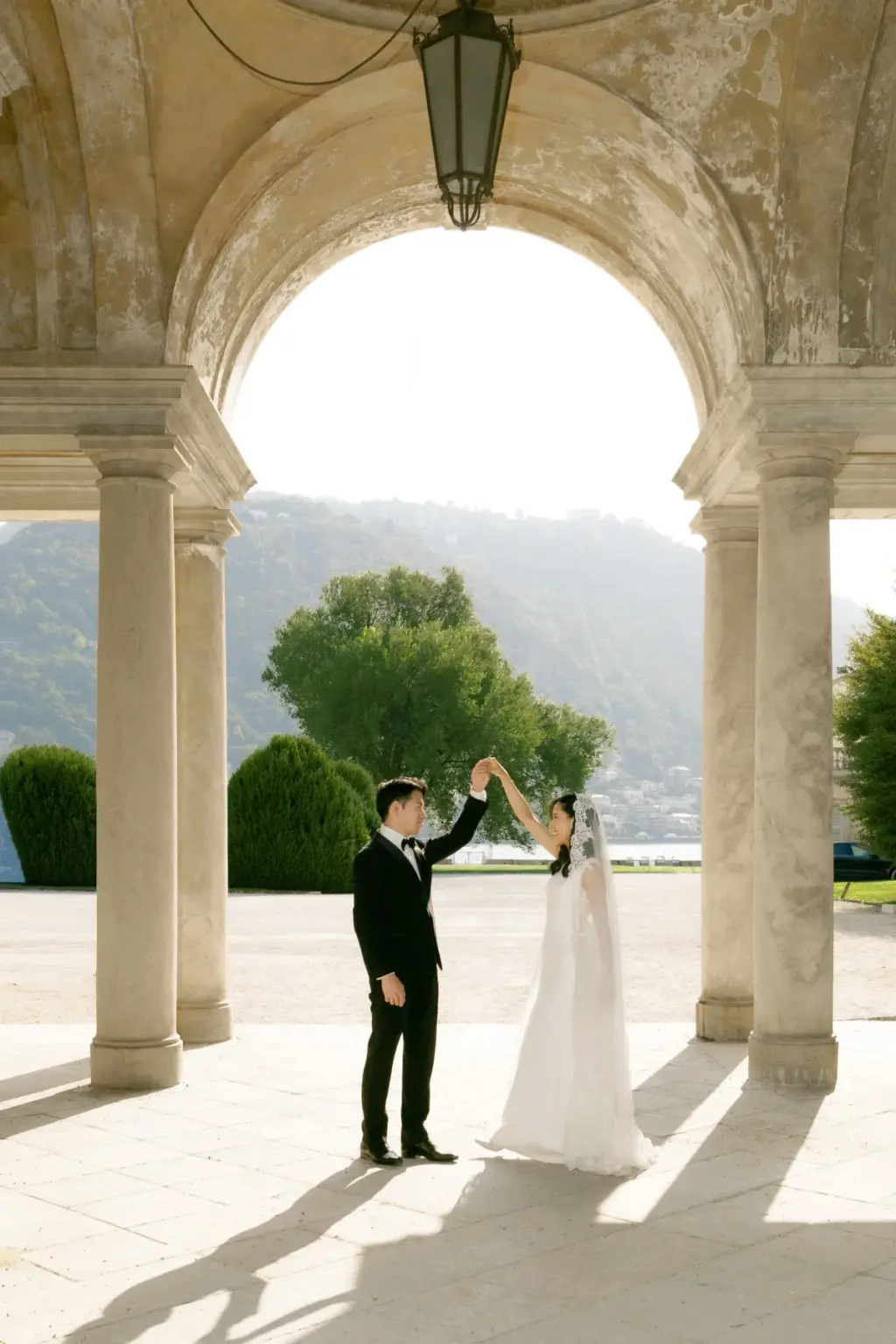 Under a stone arcade with a hanging lantern, a groom twirls his bride, Lake Como and the green hills framed by the arch.