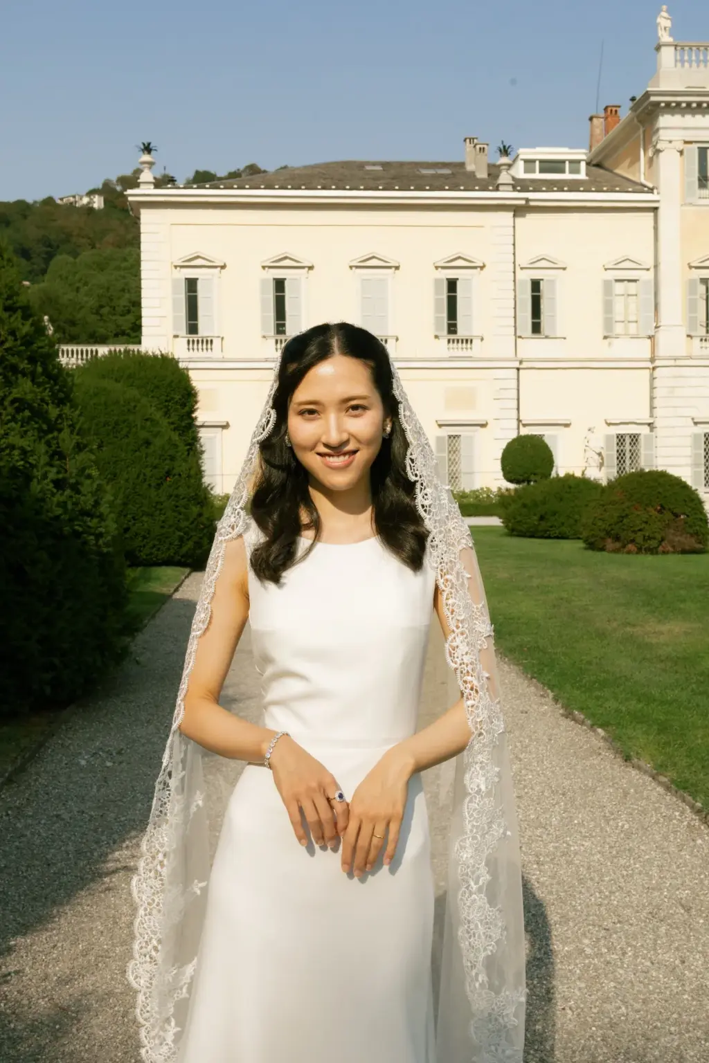 A smiling bride in a sleeveless crepe gown and lace-edged veil stands on a garden path with the white Villa Olmo behind her.