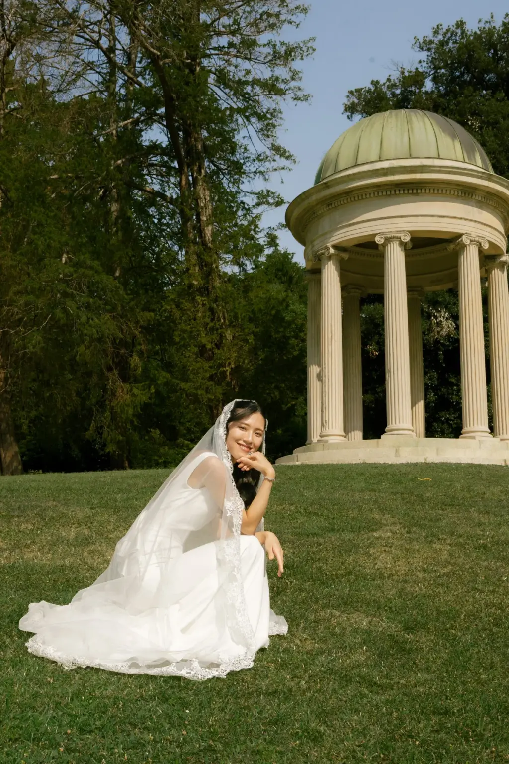 Smiling with her chin on her hand, a bride sits in her white gown on the lawn below the domed temple in the Villa Olmo park.