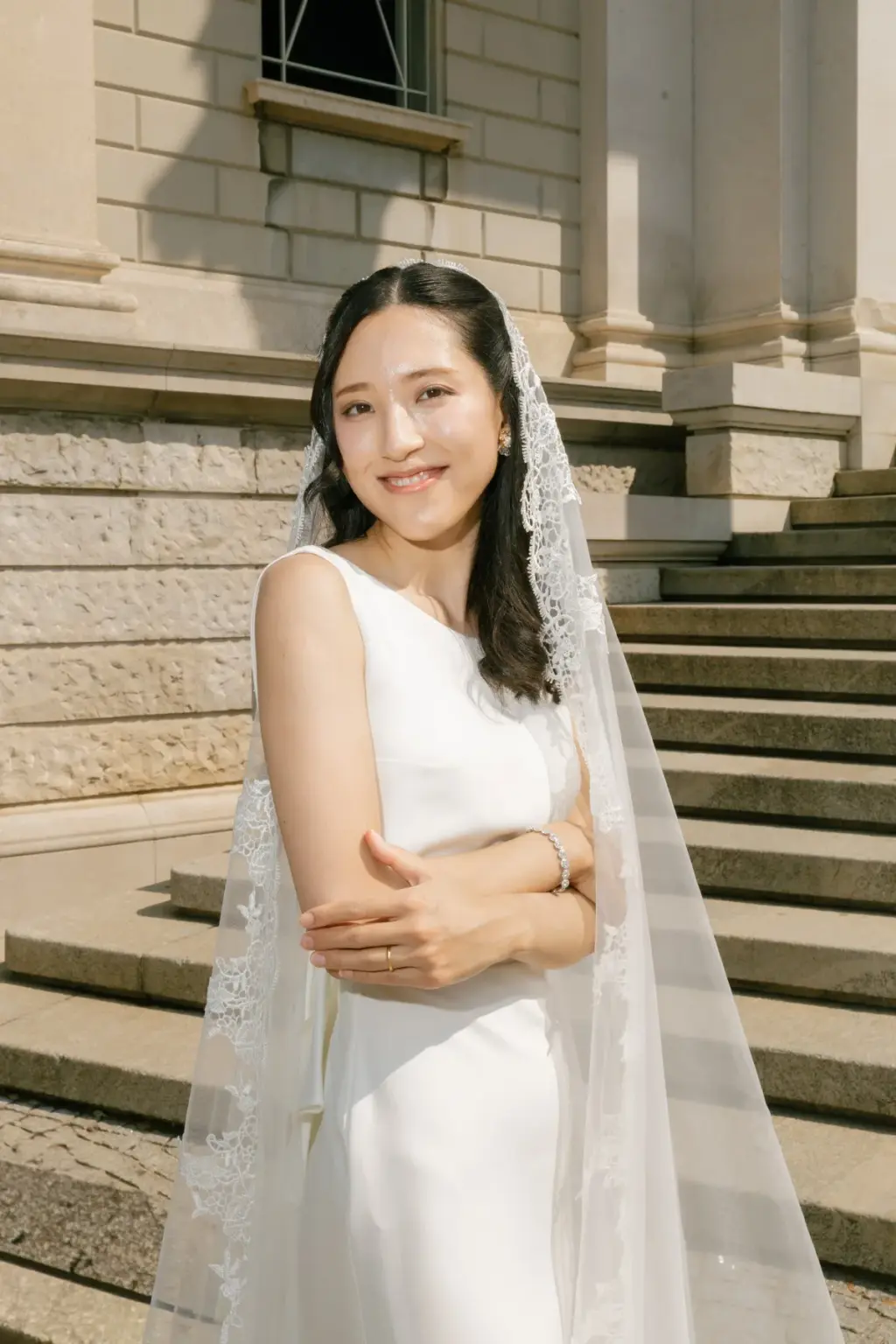 A bride with folded arms smiles on the stone steps of Villa Olmo, her long lace-edged veil falling over her shoulders.