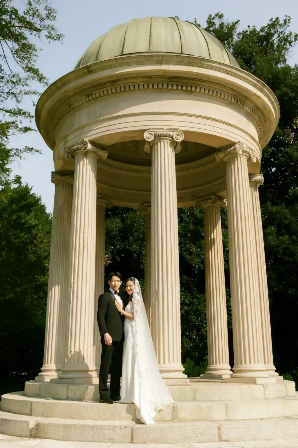 A bride and groom stand together on the steps of the round colonnaded temple in the Villa Olmo gardens, its green dome above.