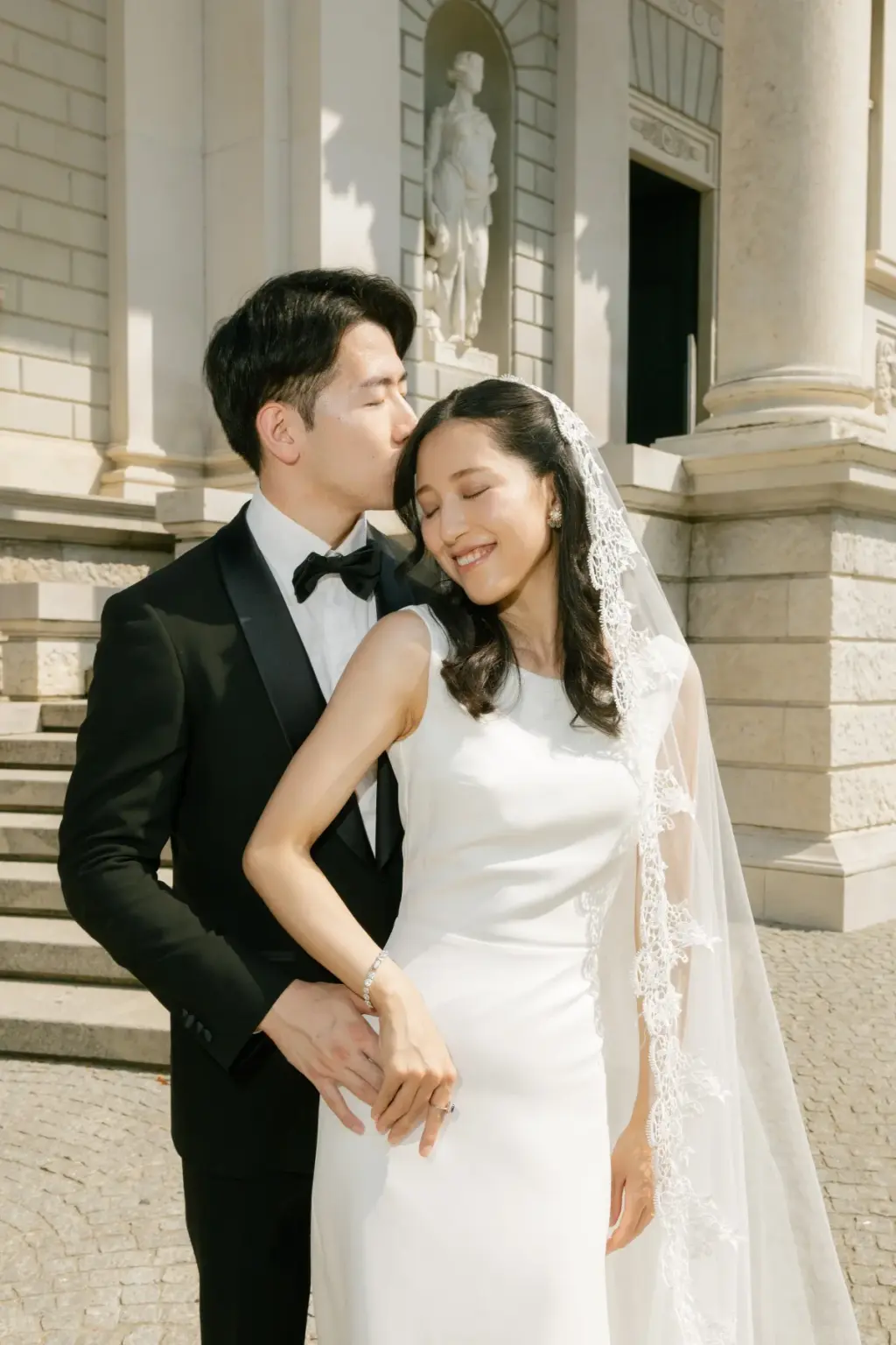 Eyes closed, a bride smiles as her groom kisses her hair from behind, his hand at her waist, by the facade of Villa Olmo.