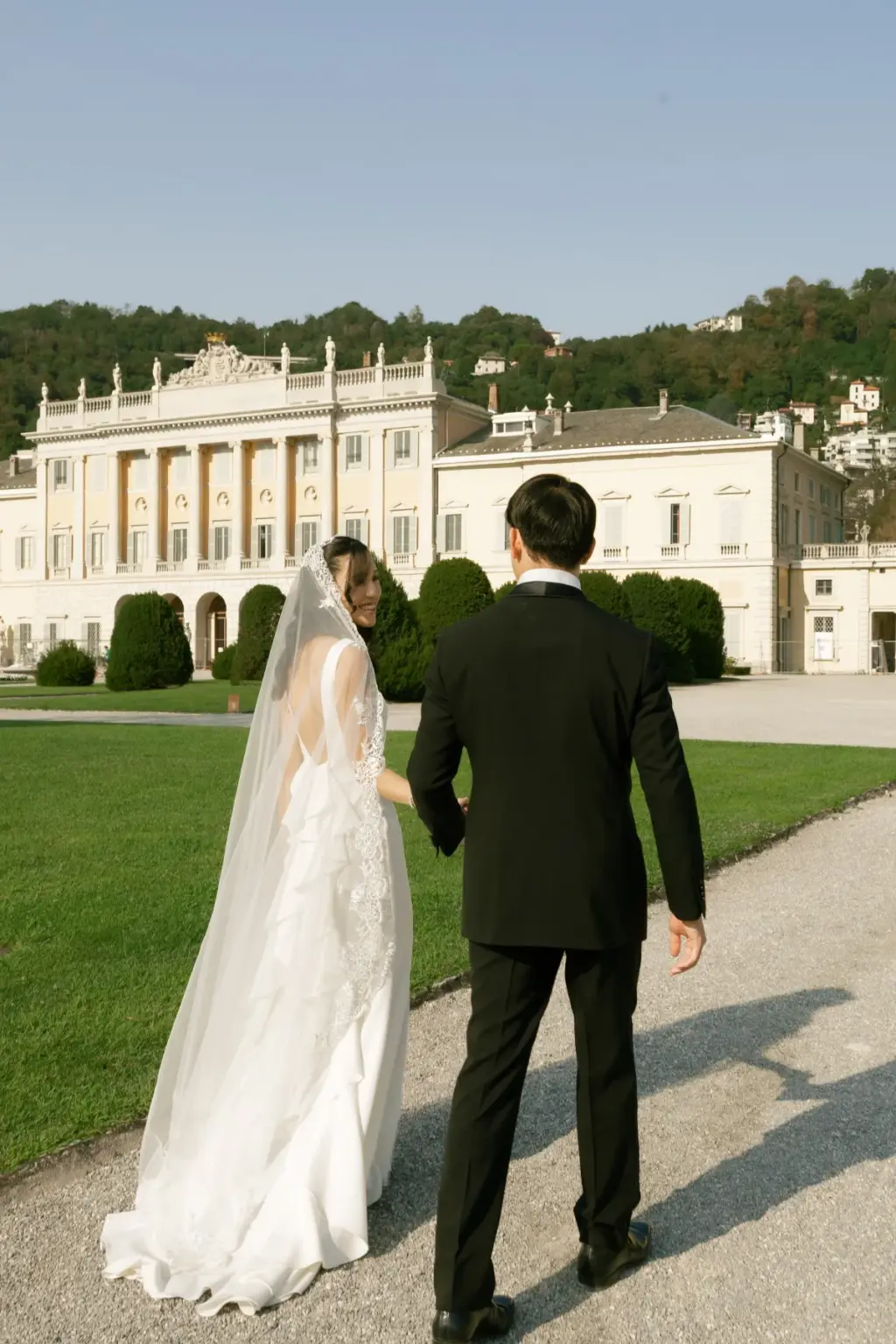 Hand in hand, a bride in a long veil and her groom walk across the lawn toward the white neoclassical facade of Villa Olmo.