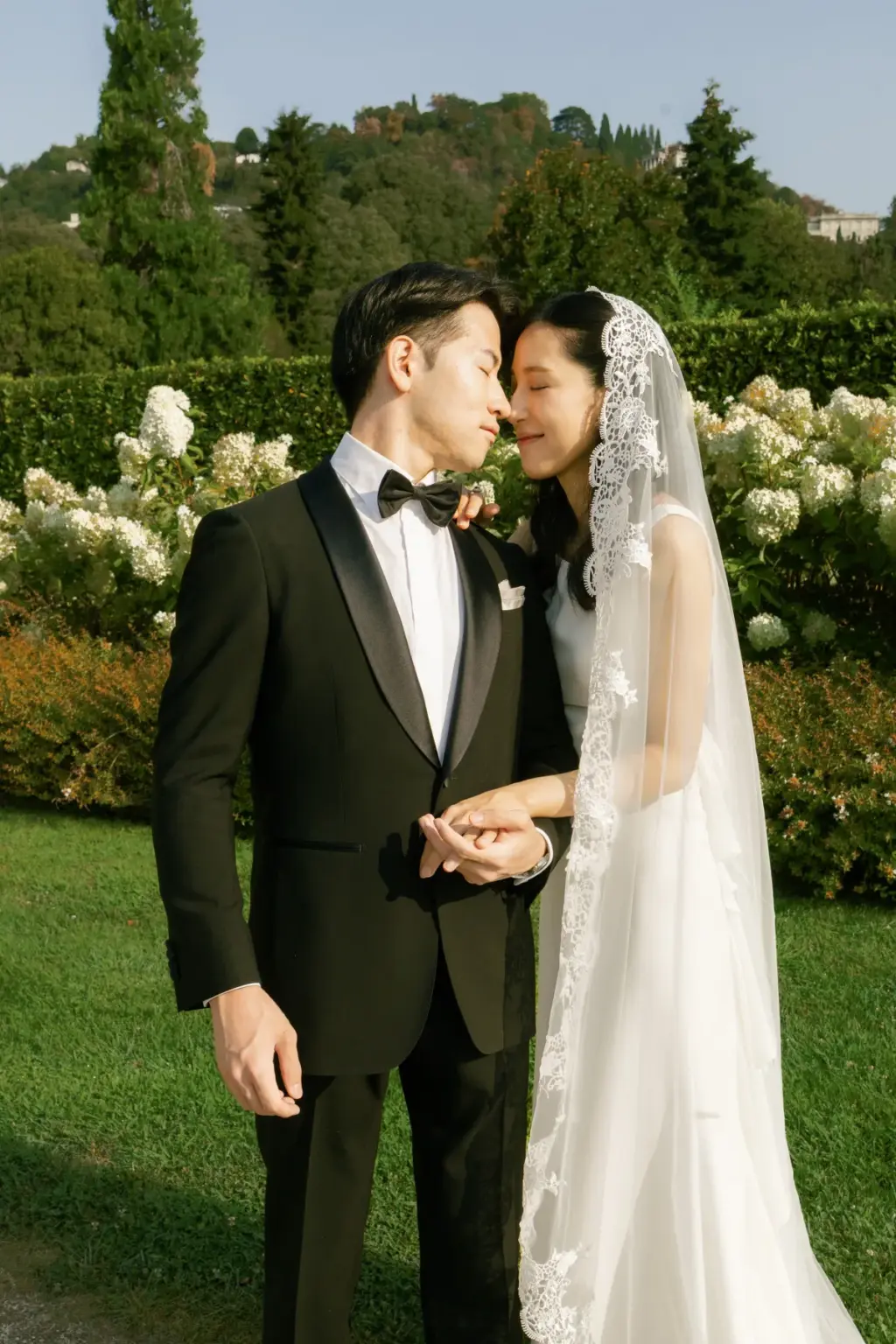 A groom in a bow tie holds his bride's hand as they lean nose to nose before white hydrangeas in the Villa Olmo gardens.