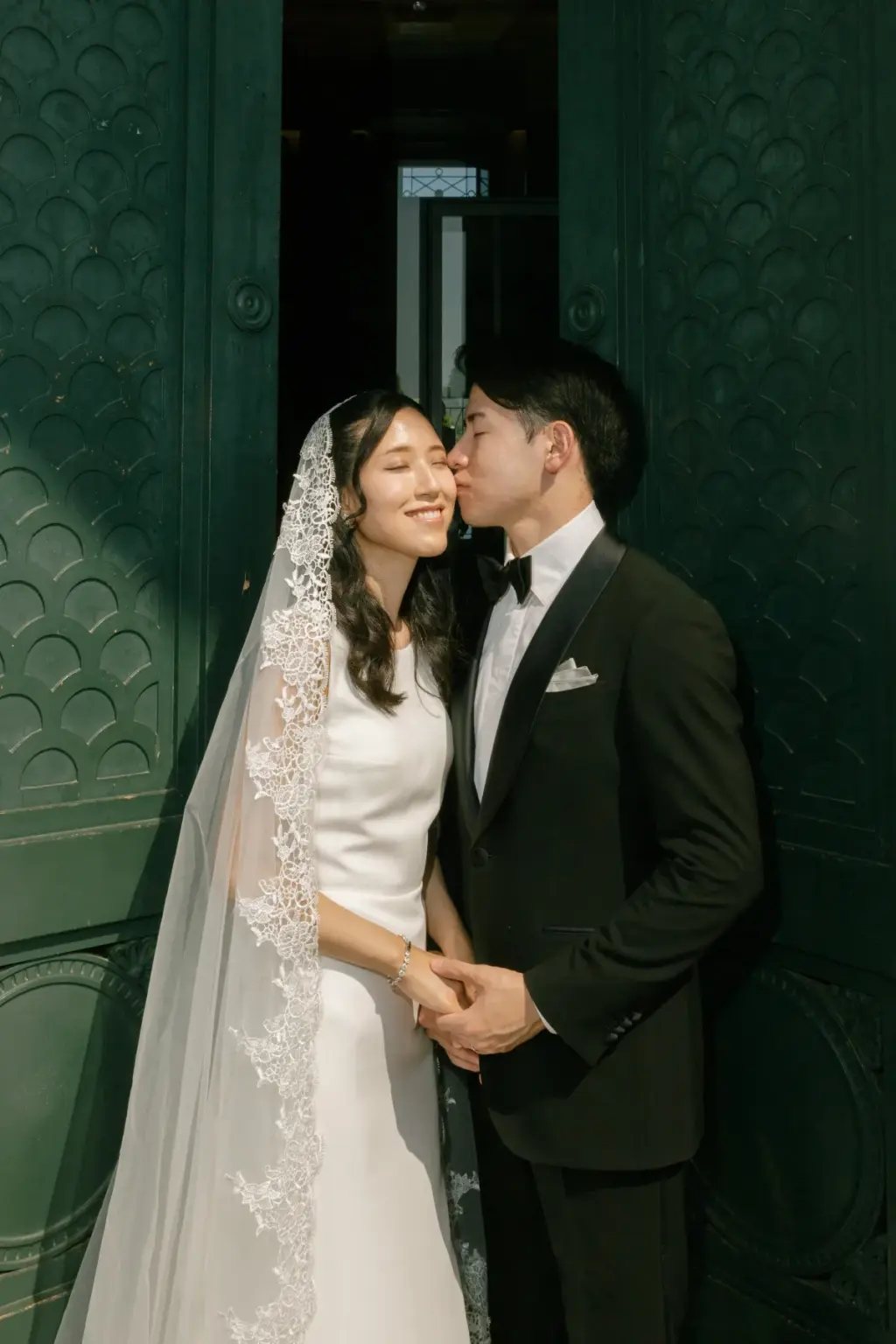 A groom kisses his smiling bride on the cheek as they hold hands in front of tall dark green panelled doors.