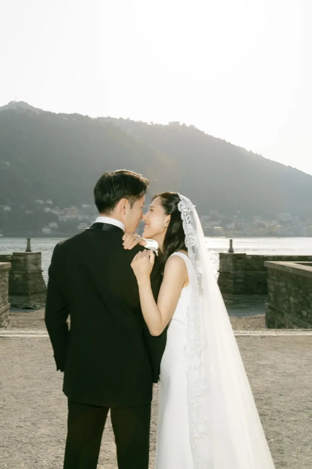 A groom in a tuxedo leans close to his bride in a lace-edged veil, noses almost touching, on the Lake Como promenade at Villa Olmo.