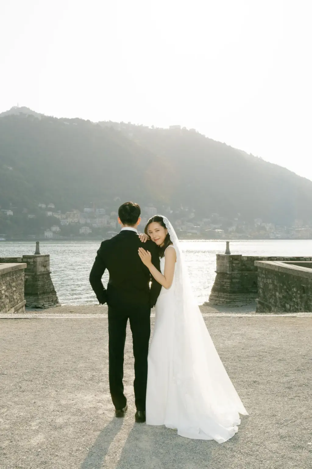 A bride rests her hands on her groom's shoulder and smiles back over it as they face Lake Como from the Villa Olmo waterfront.