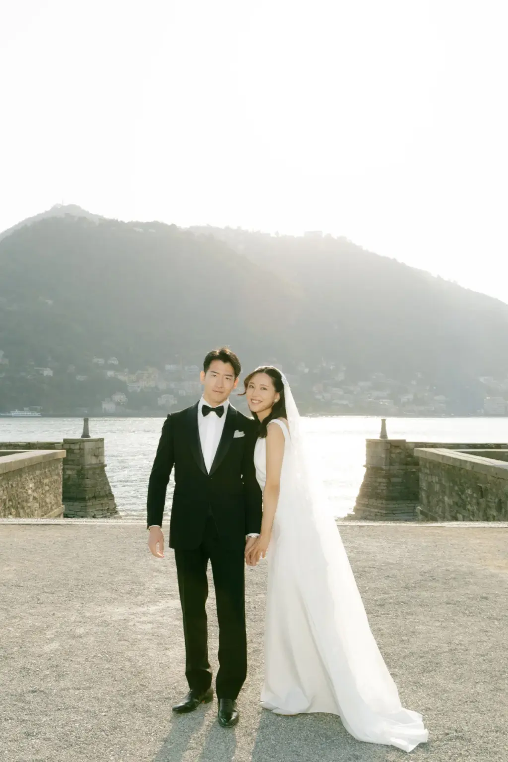 A bride in a long veil and a groom in a black tuxedo stand side by side on the lakefront at Villa Olmo, the Como hills behind.