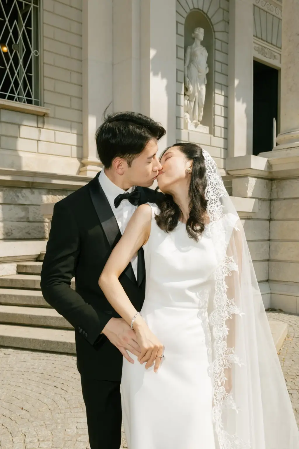 A bride and groom kiss on the stone steps of Villa Olmo, a statue in a niche on the pale facade above them.