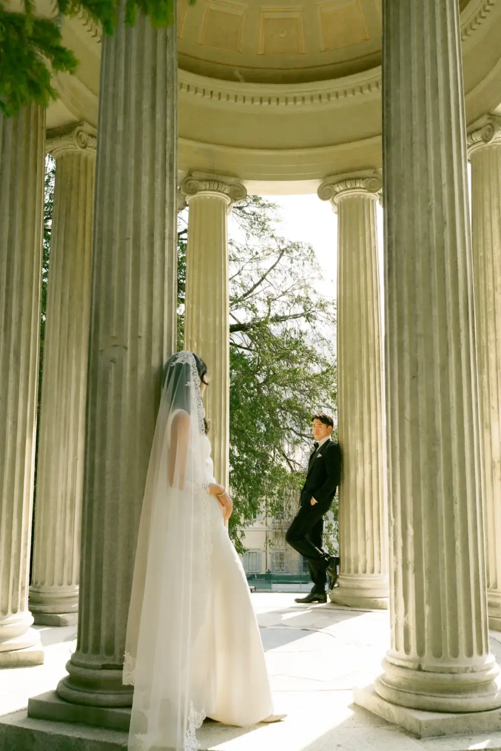 A bride in a long veil leans on a fluted column while her groom stands across from her inside the round temple at Villa Olmo.