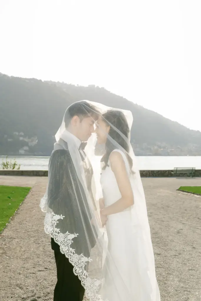 A bride and groom touch foreheads under her lace-trimmed veil on a gravel path by Lake Como in the Villa Olmo gardens.