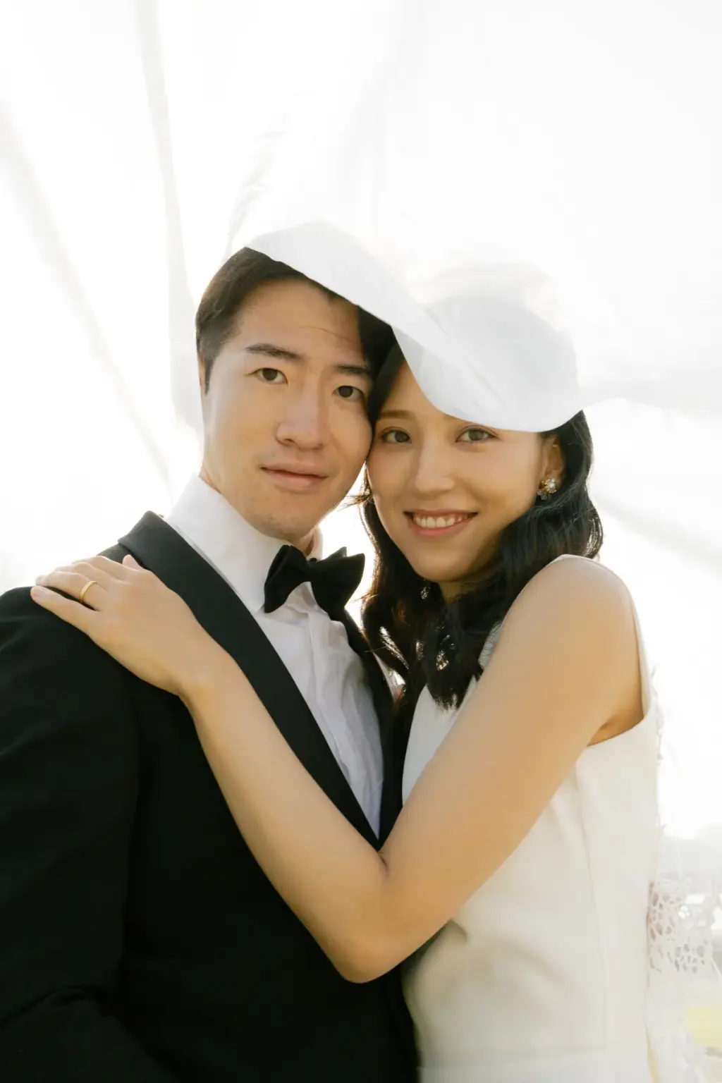 Close portrait of a smiling bride and groom wrapped in the soft white glow of her veil, her arm around his shoulders.